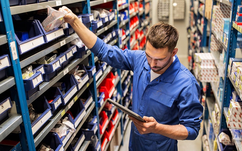 A man uses a tablet while retrieving an item from shelves of parts bins in a warehouse.
